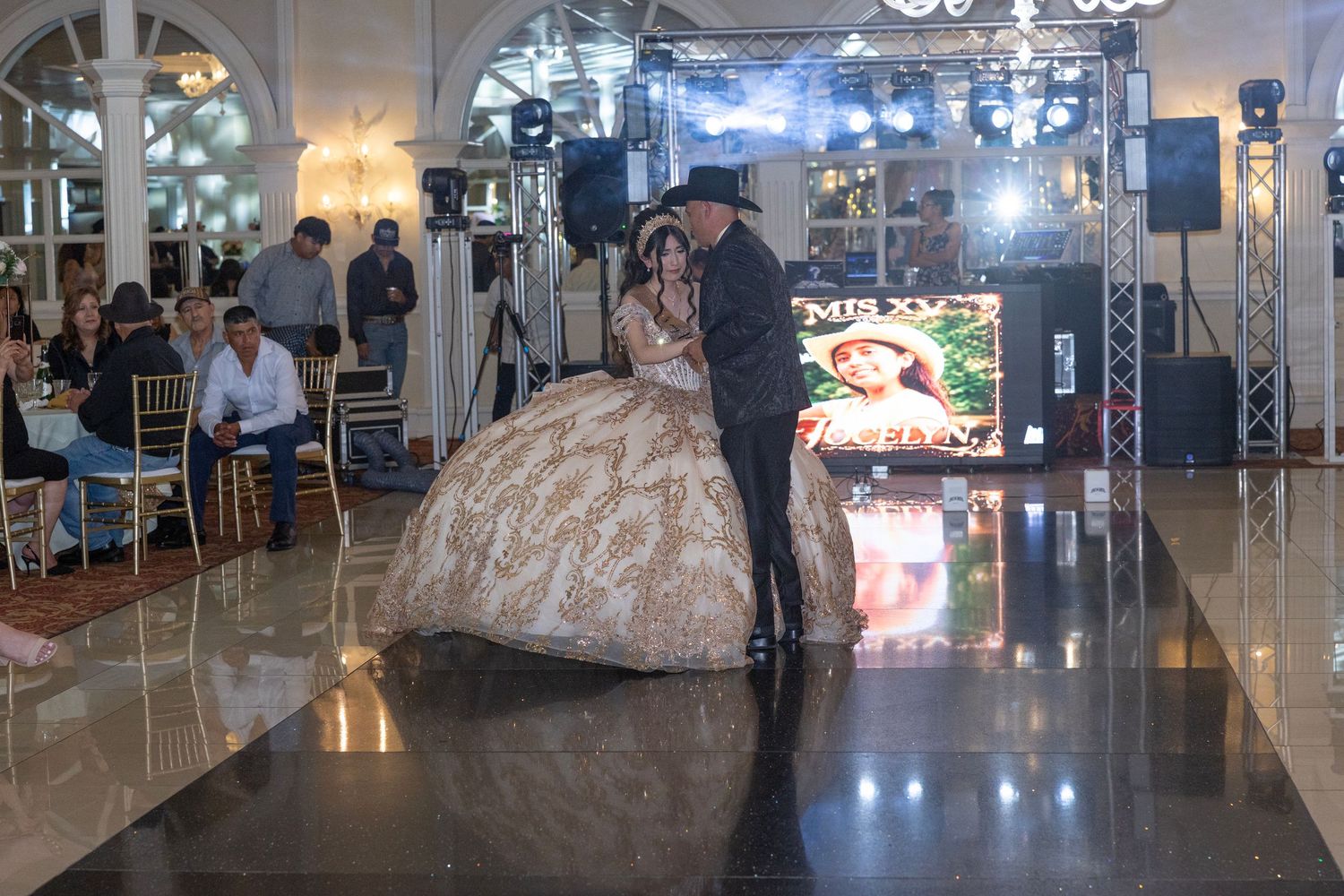 A couple shares a dance at an elegant quinceañera celebration in a grand ballroom with guests seated nearby.