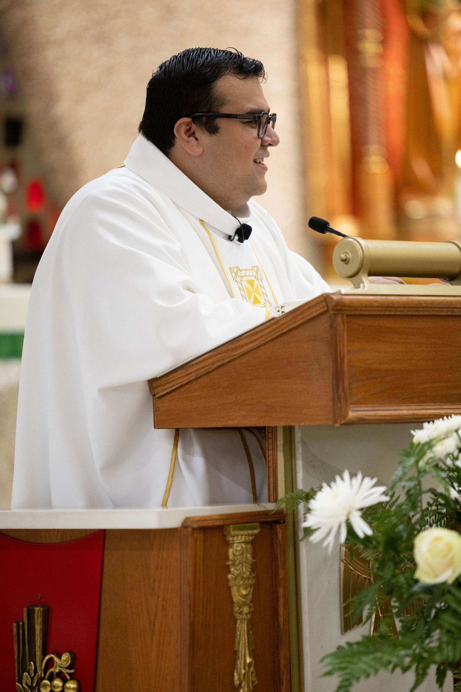 Catholic priest in white vestments smiling at pulpit during Mass ceremony in church.