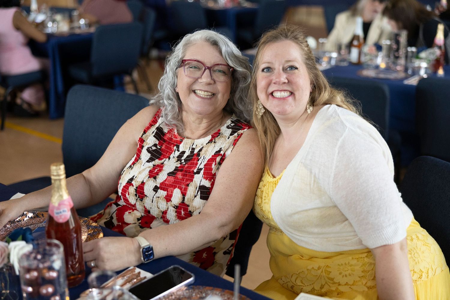Two smiling women pose together at a formal banquet event, one in a floral dress and one in yellow.