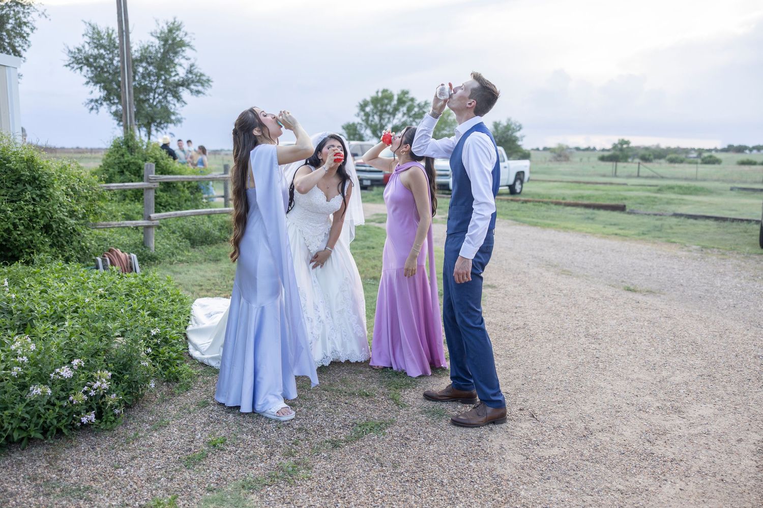 Four people in formal attire, including wedding dresses, celebrate outdoors on a gravel path at a rustic farm venue.