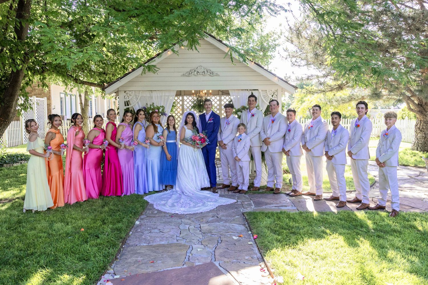Wedding party poses outdoors by white gazebo, bridesmaids in colorful gowns, groomsmen in gray suits.
