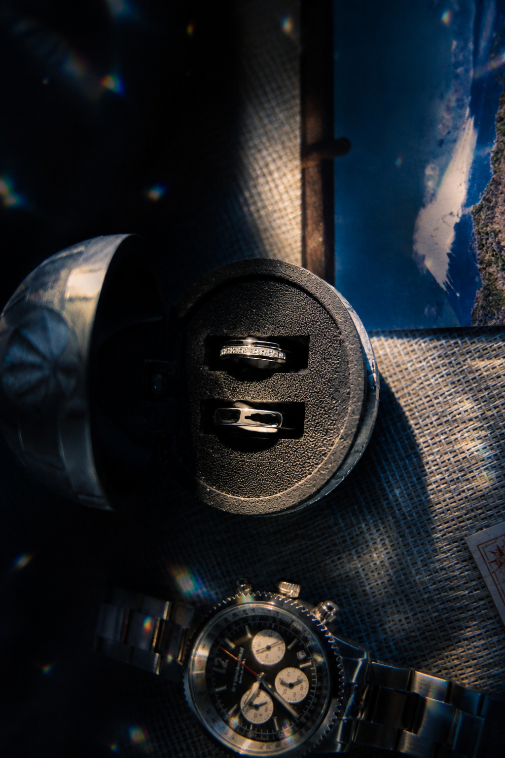 Two wedding rings displayed on a black jewelry box beside a luxury chronograph watch in dramatic dark lighting.