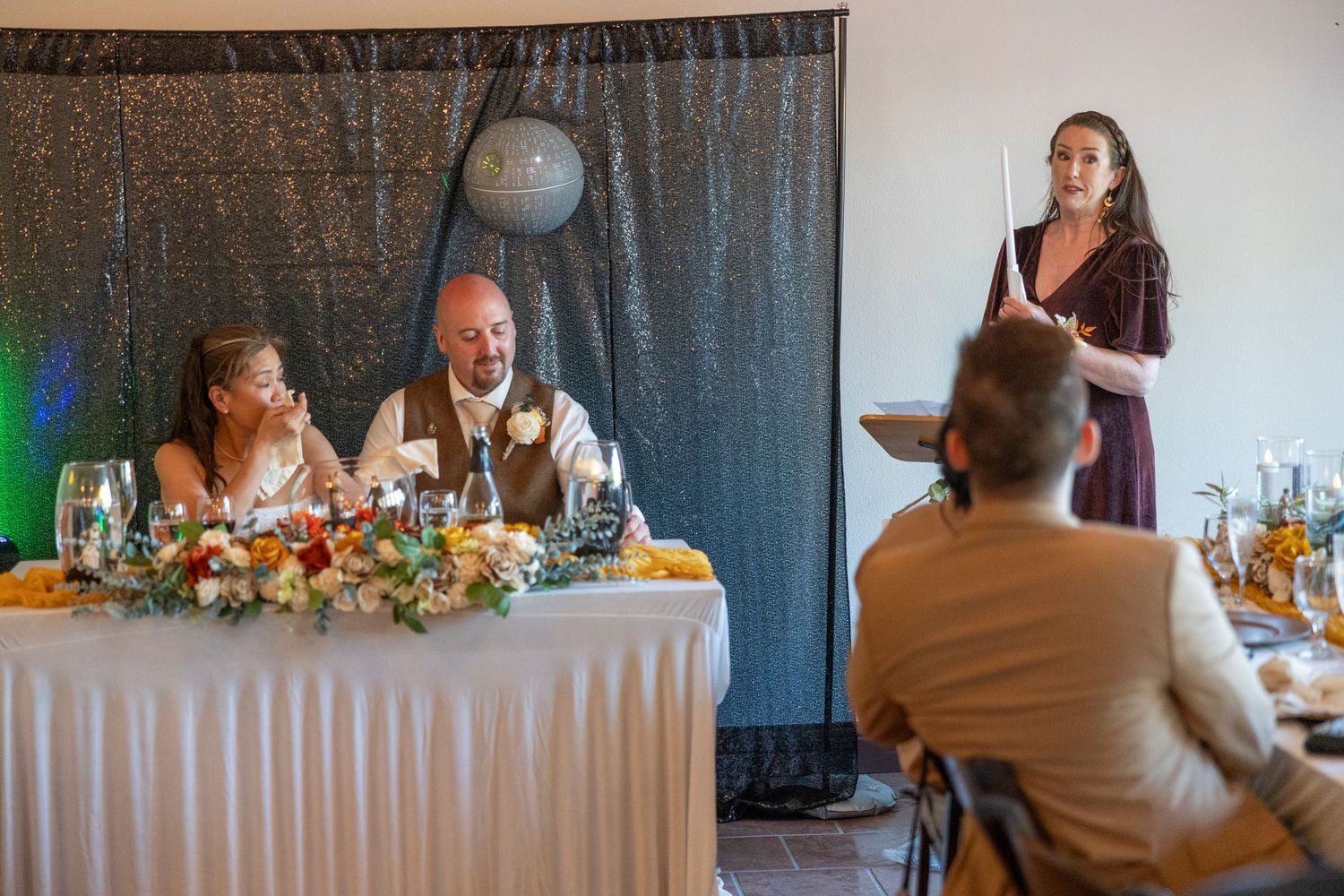 A woman gives a toast speech at a wedding reception while the bride and groom smile at a flower-adorned head table.