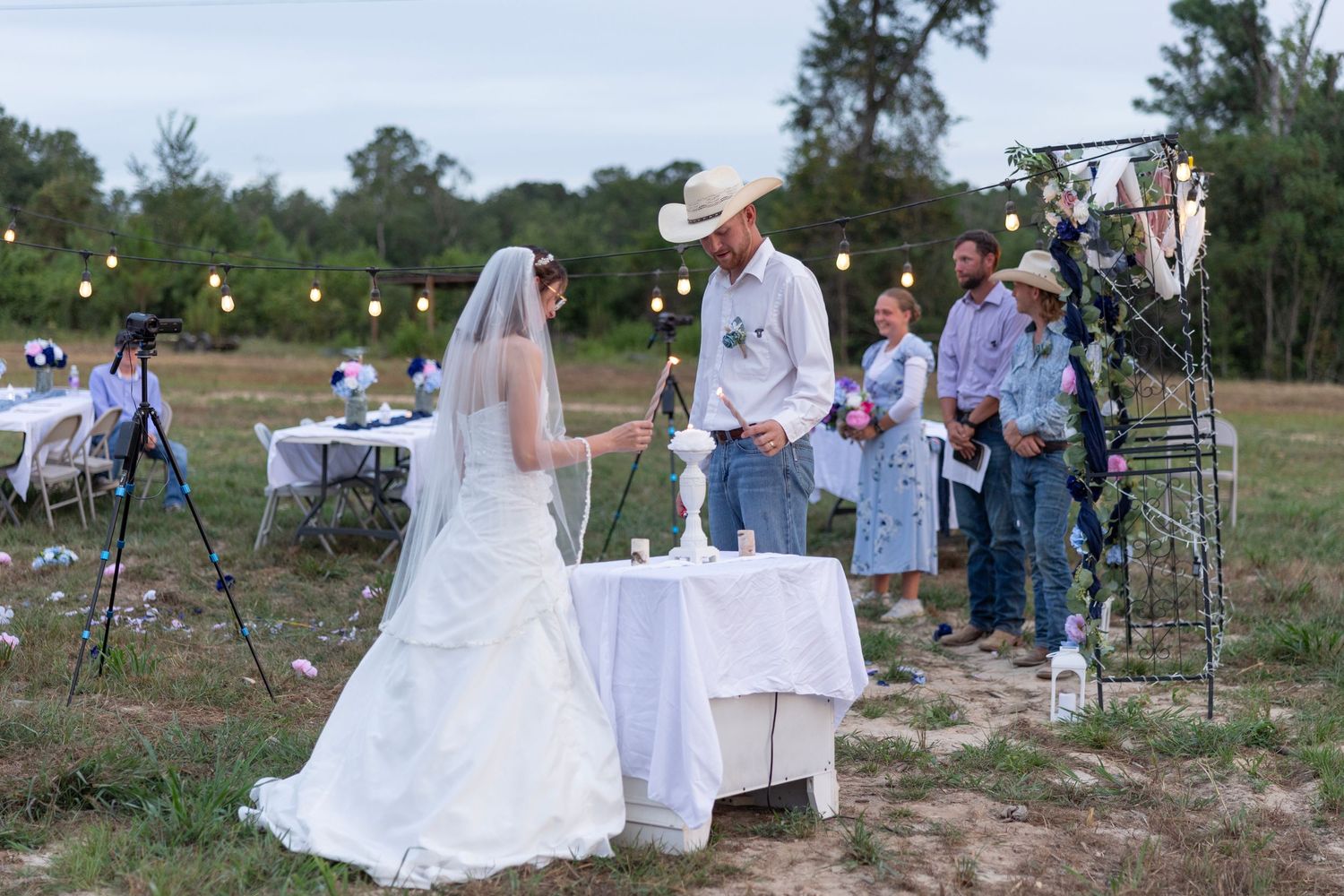 Outdoor rustic wedding ceremony in a field with bride in white gown and groom in cowboy hat at decorated altar.