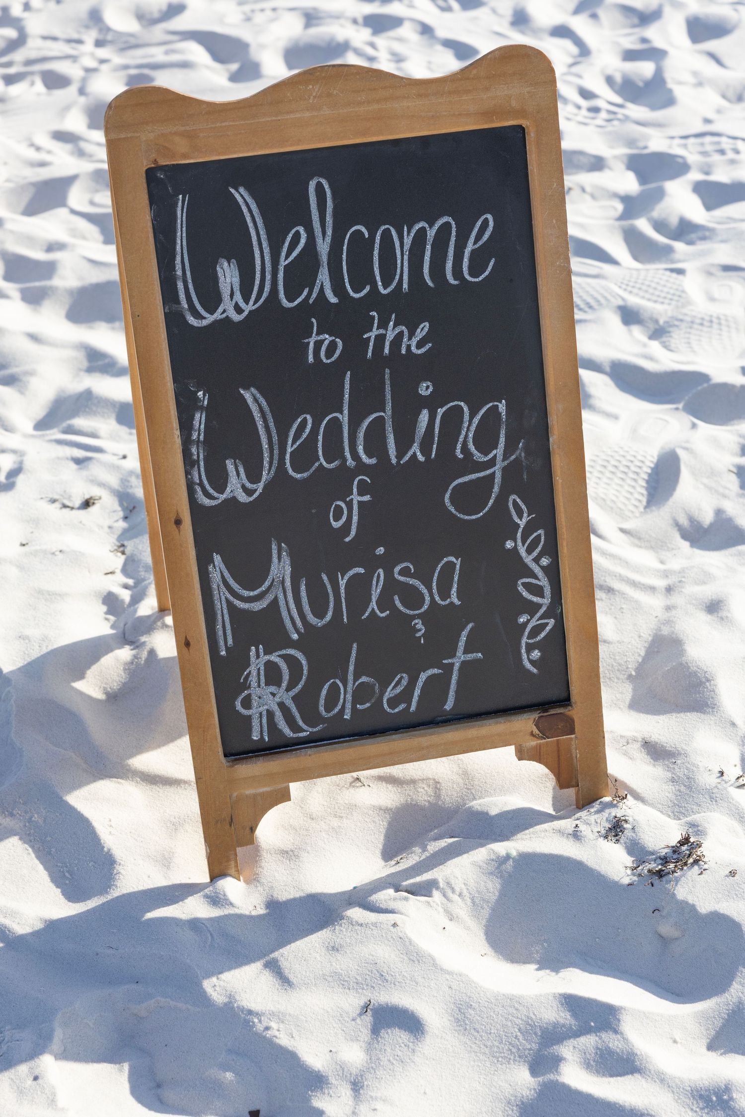 Chalkboard wedding welcome sign for Murisa and Robert displayed in white sand at a beach wedding.