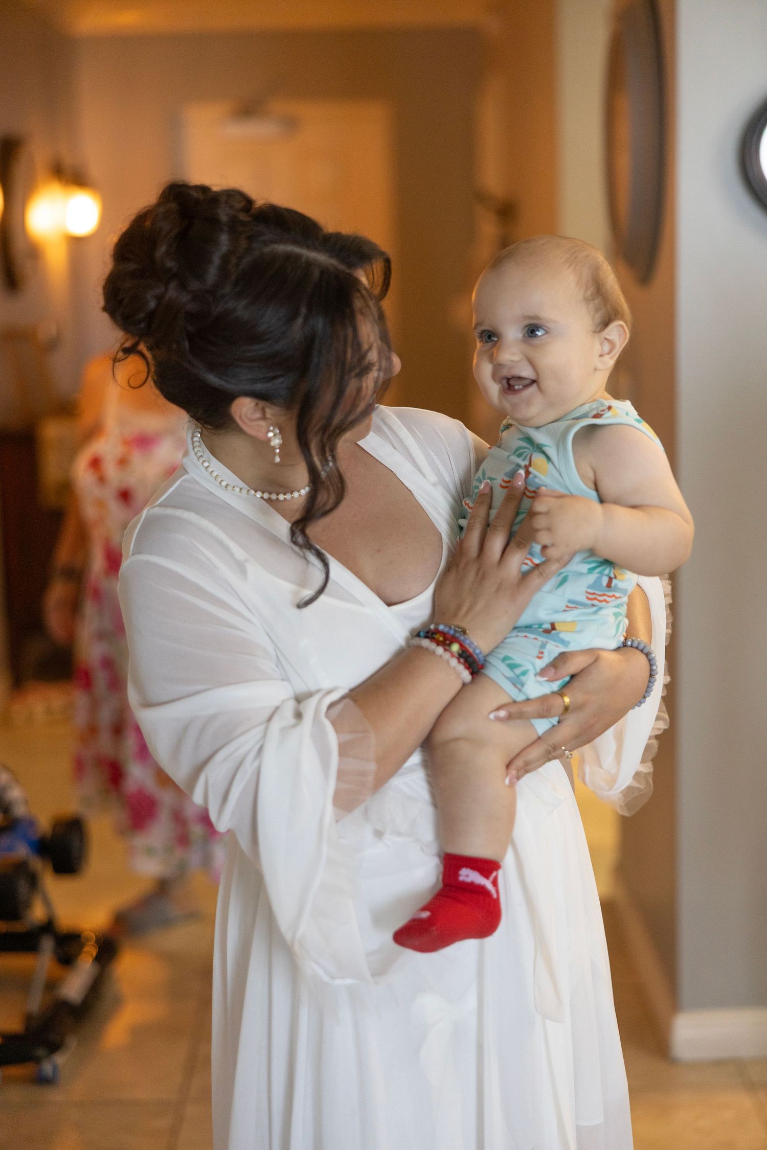 Bride in white dress holding a smiling baby in blue romper with red Puma socks at a wedding venue.