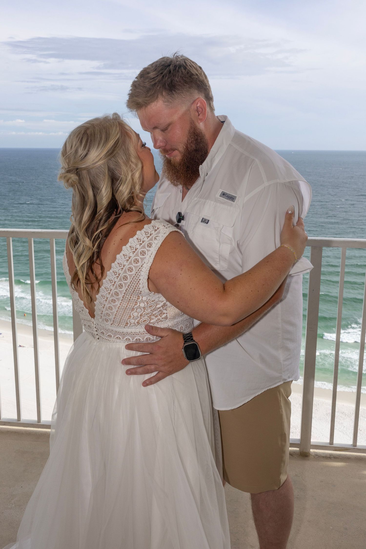Couple sharing romantic kiss in wedding attire on oceanfront balcony with white sand beach and blue water backdrop.