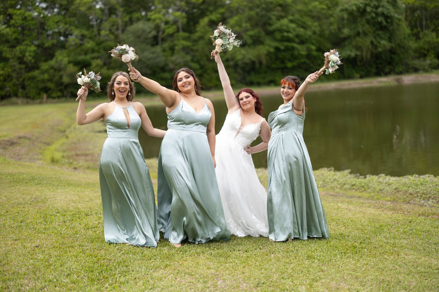 A bridal party in mint green bridesmaid dresses and white wedding gown pose joyfully by a pond while holding bouquets.