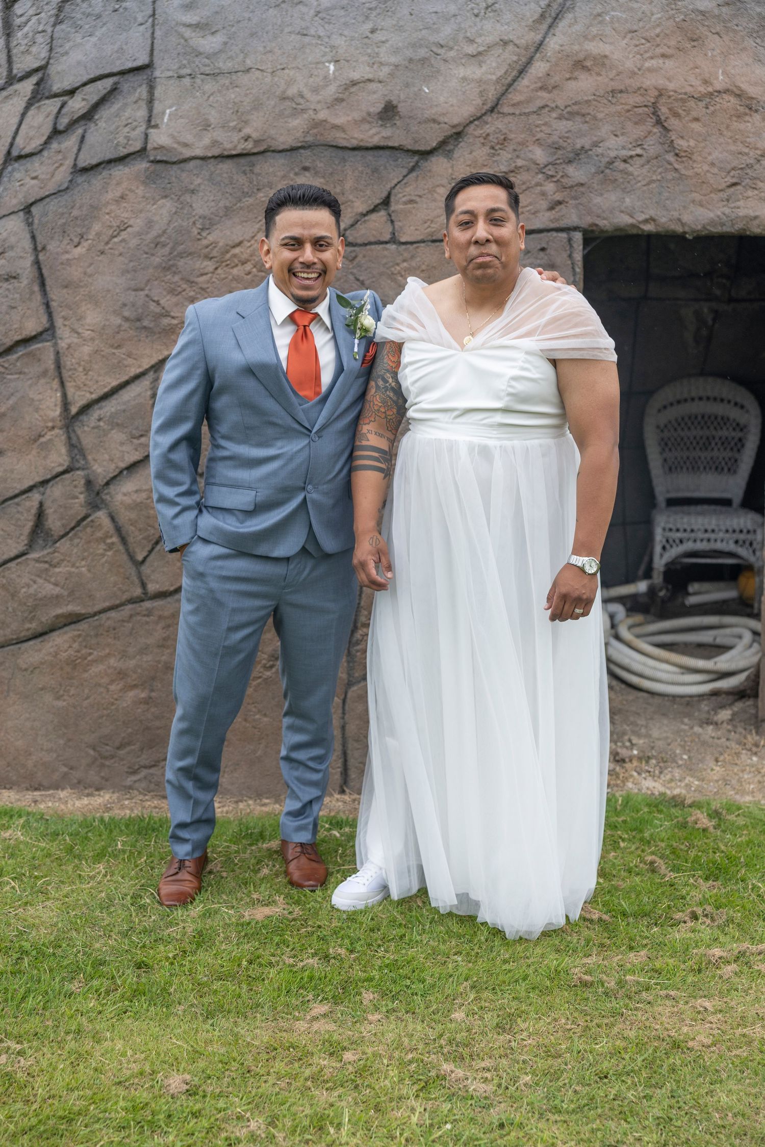 A couple in wedding attire stands together on grass against a stone wall background.