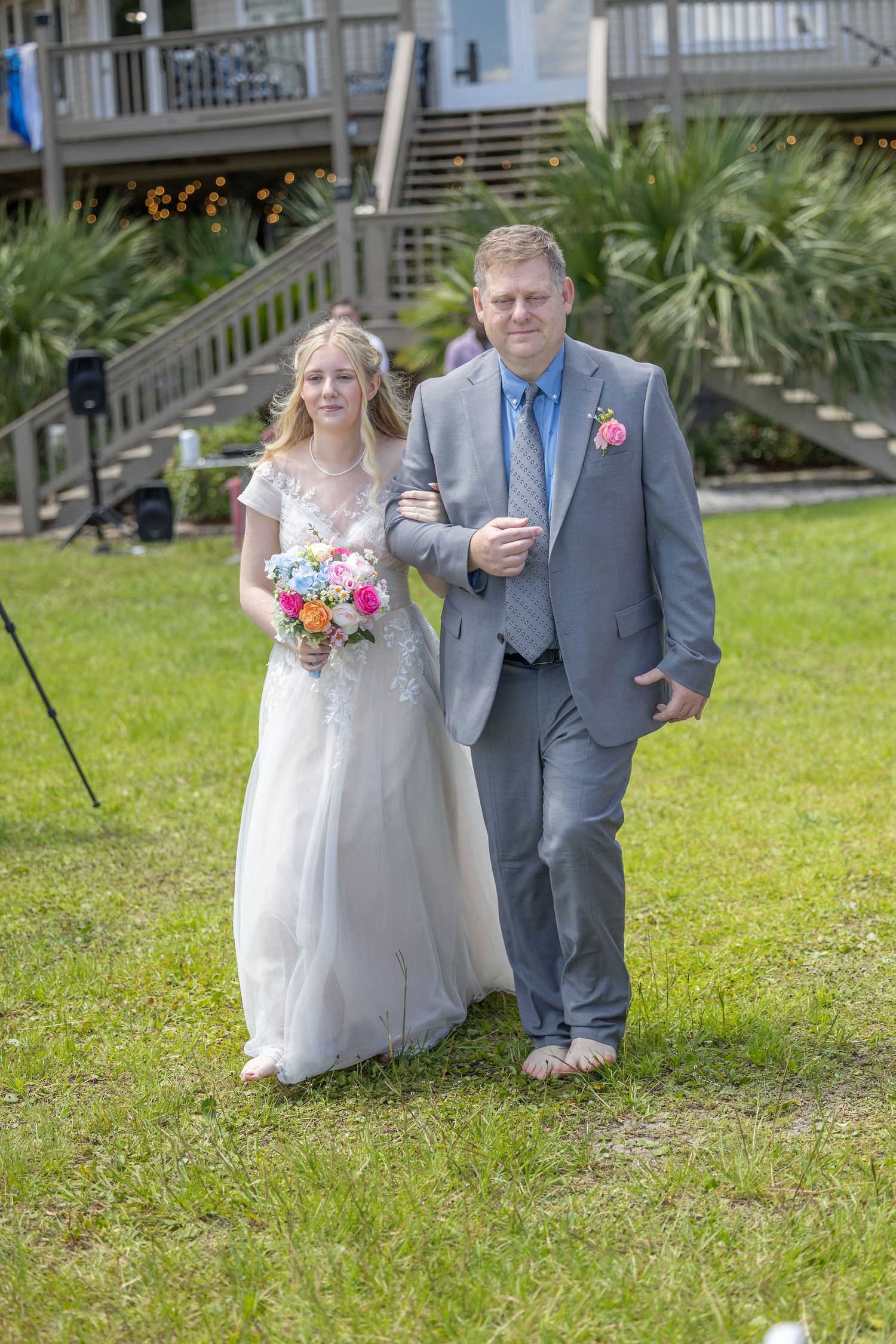 A couple walks arm in arm on a grassy lawn outside a house with wooden stairs and palm trees in the background.