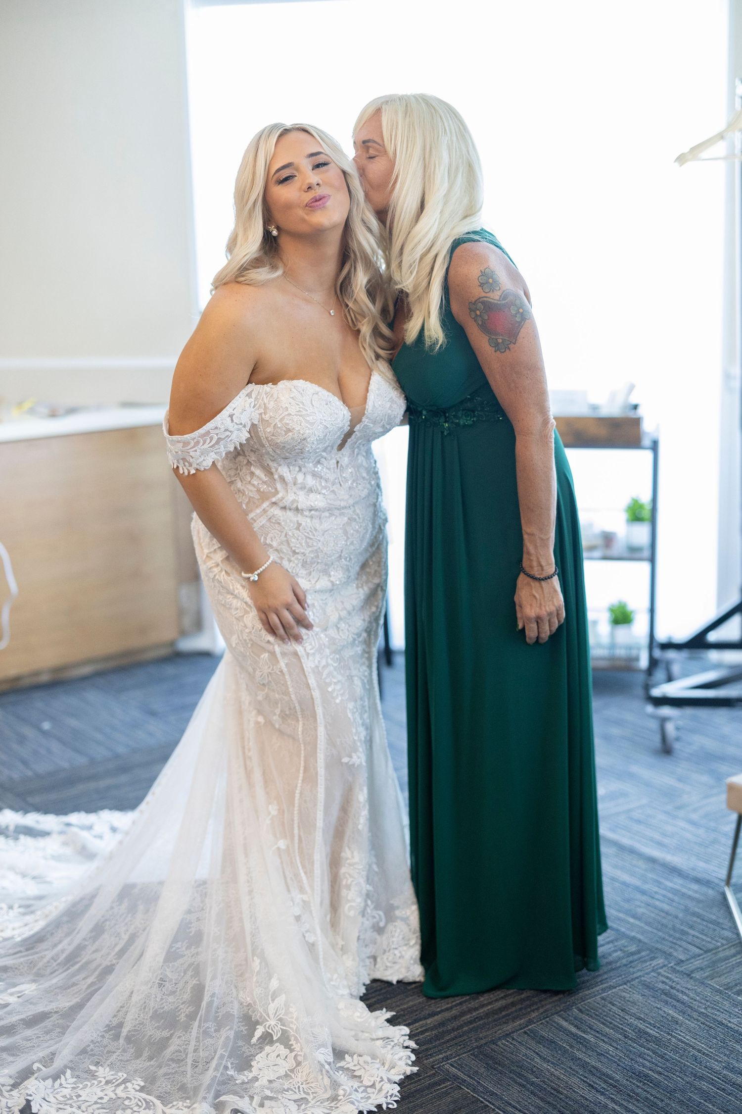 Two women pose together indoors, one wearing a white lace wedding dress and the other in an emerald green formal gown.