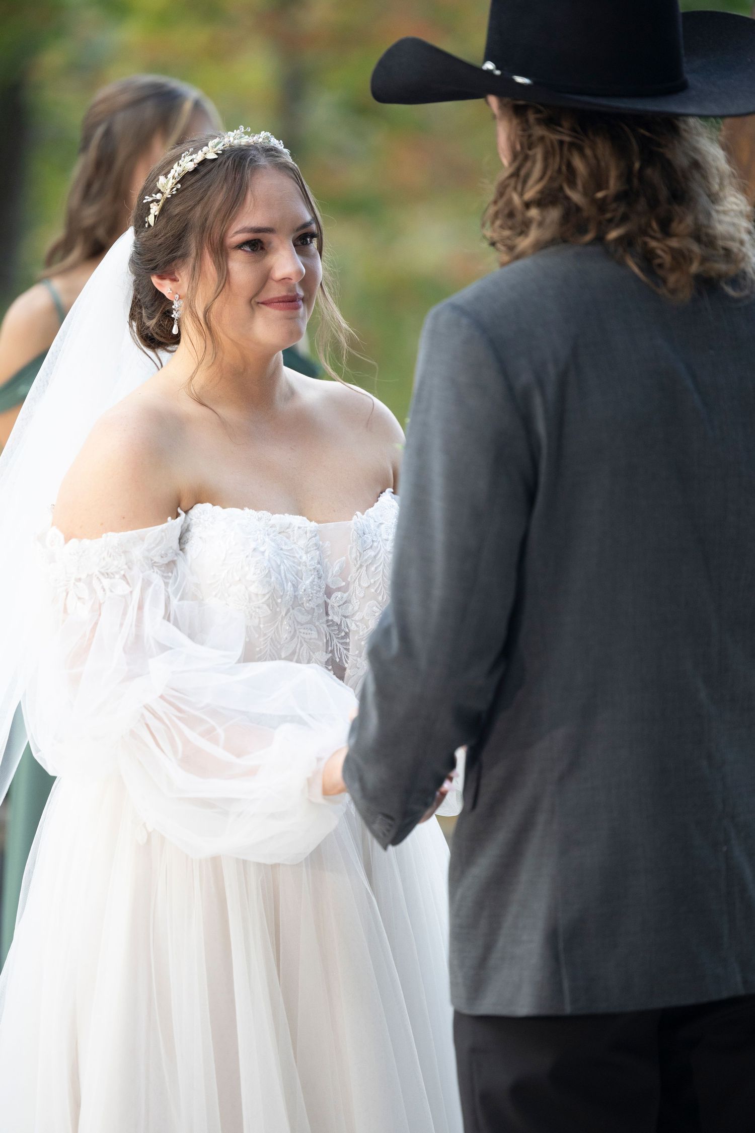 A romantic outdoor wedding ceremony shows a bride in a flowing off-shoulder white gown facing someone wearing a black cowboy hat.