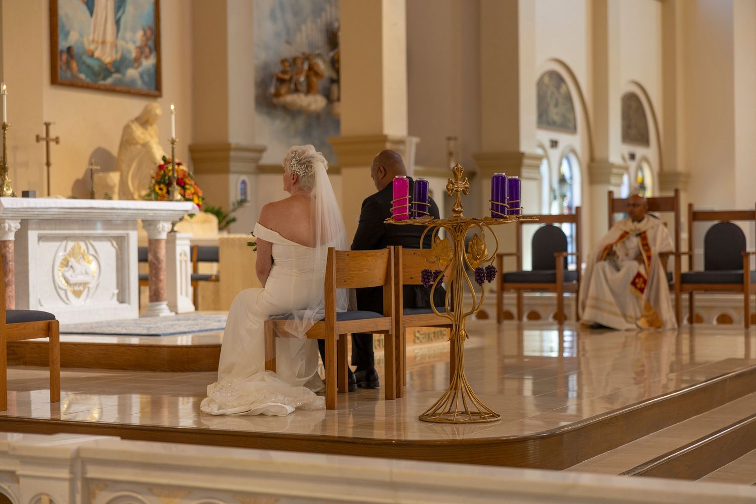 A couple sits in a Catholic church during a wedding ceremony with white marble altar and arched architecture.