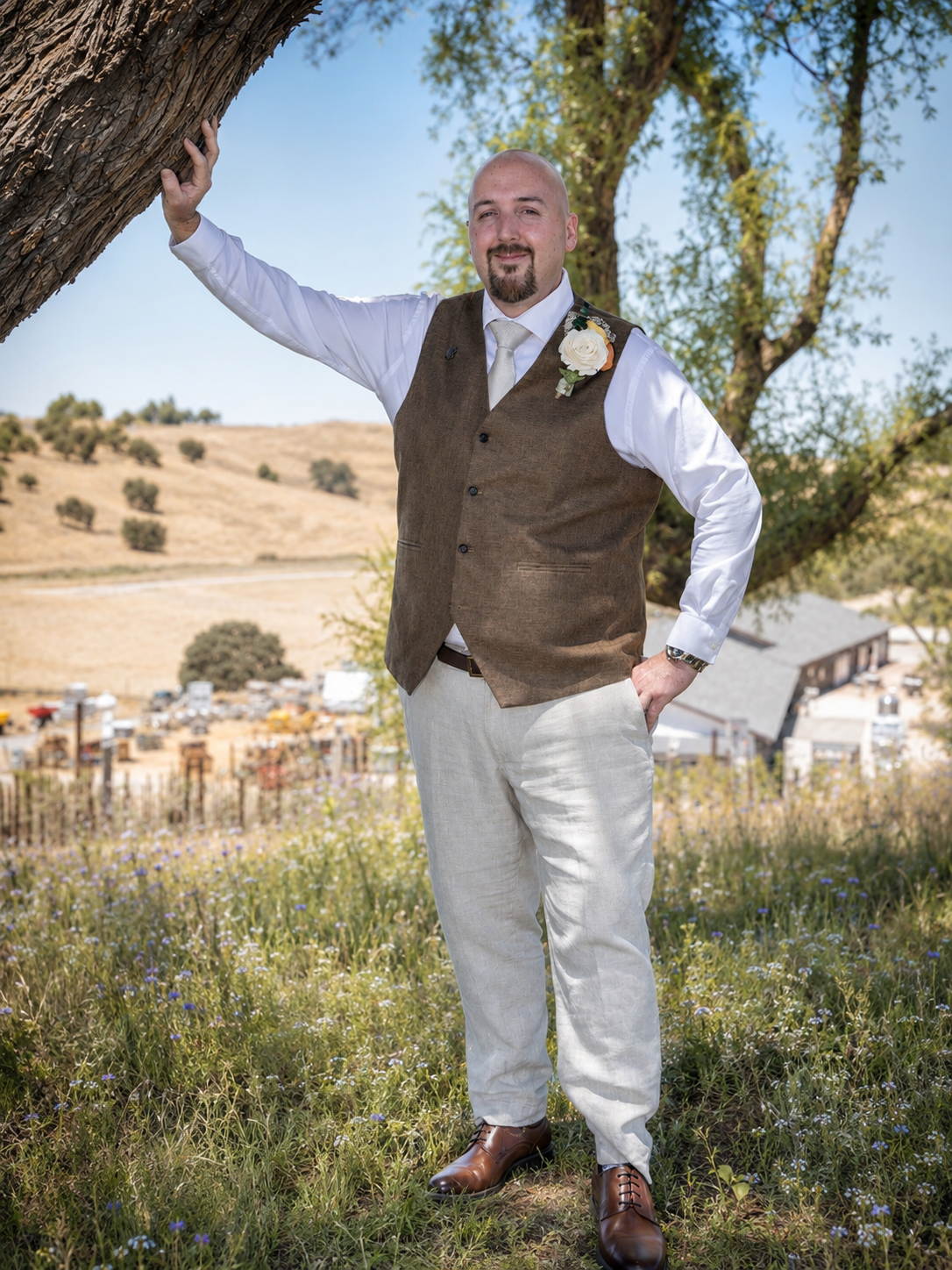 Groom in brown vest and white shirt stands by a tree in a rustic outdoor wedding setting with rolling hills.