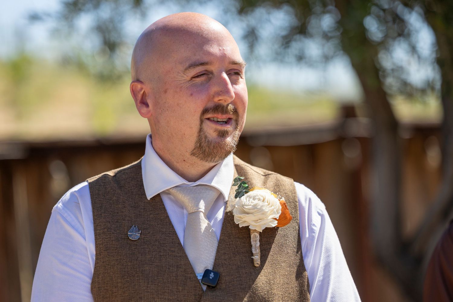 Bald groom wearing brown tweed vest with boutonniere and tie smiling outdoors at wedding ceremony.