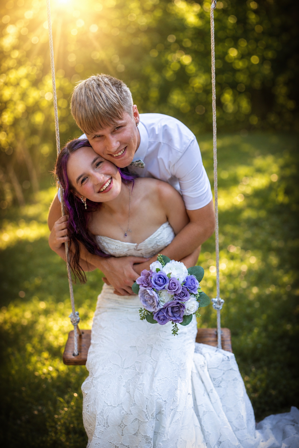 Smiling bride sits on a swing holding purple bouquet while groom embraces her in golden sunlight.