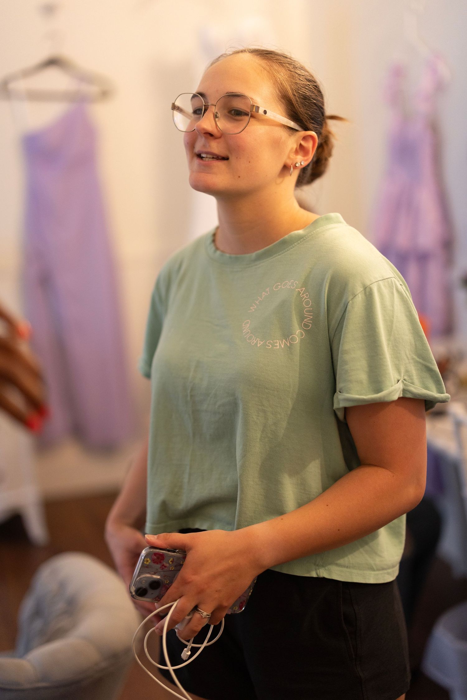 Young woman in mint green t-shirt holding a device, standing in a boutique with purple dresses in background.