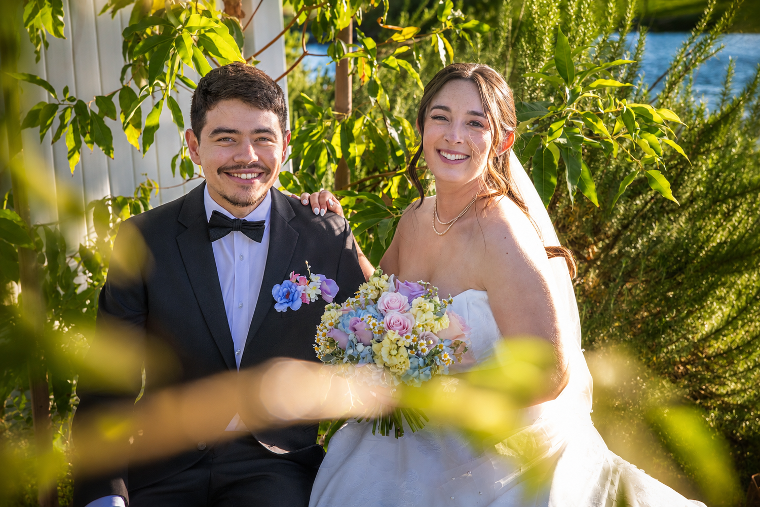 Smiling bride and groom pose among lush green foliage, bride holding colorful bouquet in white gown.