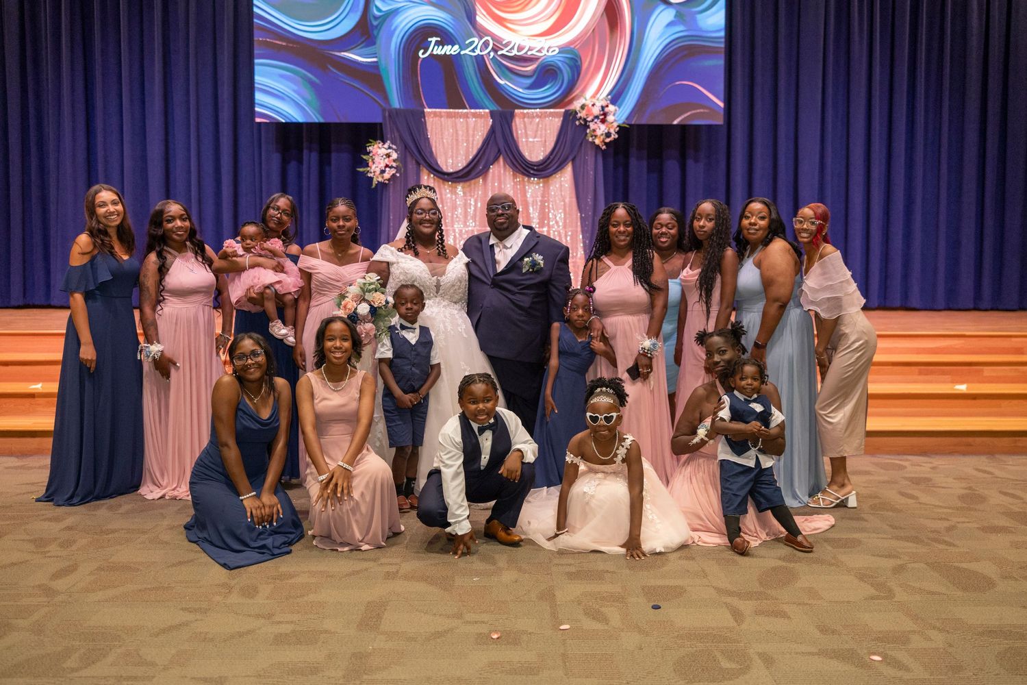 Wedding party poses together in front of a decorated stage, bride and groom surrounded by bridesmaids in pink and blue gowns.
