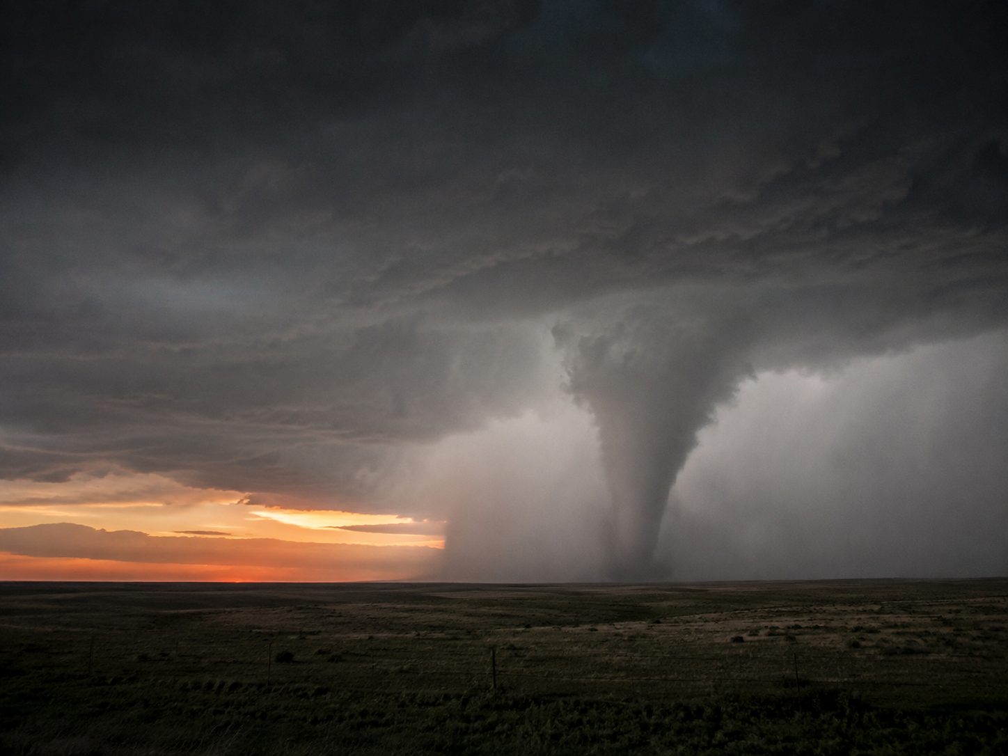 A massive tornado touches down at sunset over a flat prairie landscape under dramatic storm clouds.