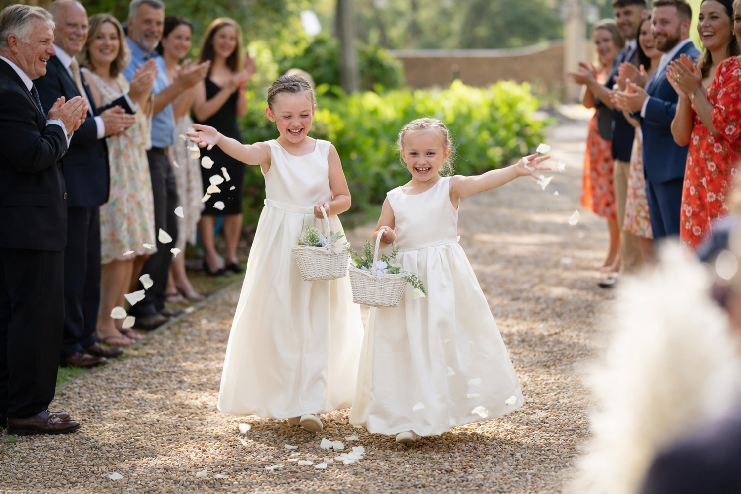 Two flower girls in white dresses toss petals down the aisle at an outdoor wedding ceremony.