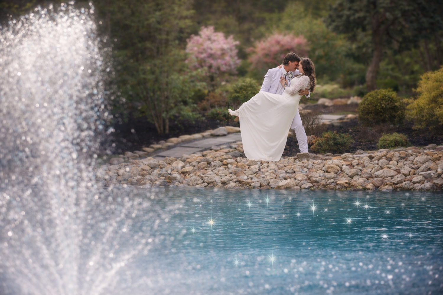 Couple embracing by a sparkling fountain pond with cherry blossoms, bride in flowing white gown lifted by groom.