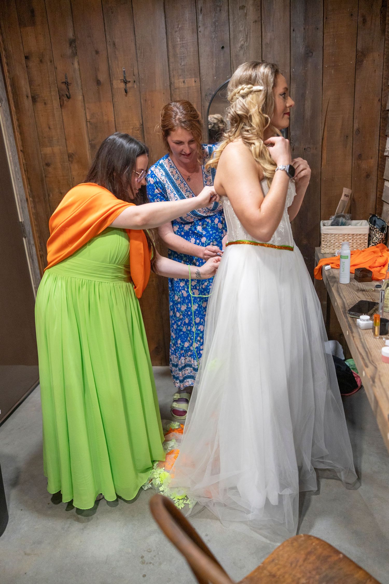 Two bridesmaids help a blonde bride into her white wedding gown in a rustic wood-paneled bridal suite.