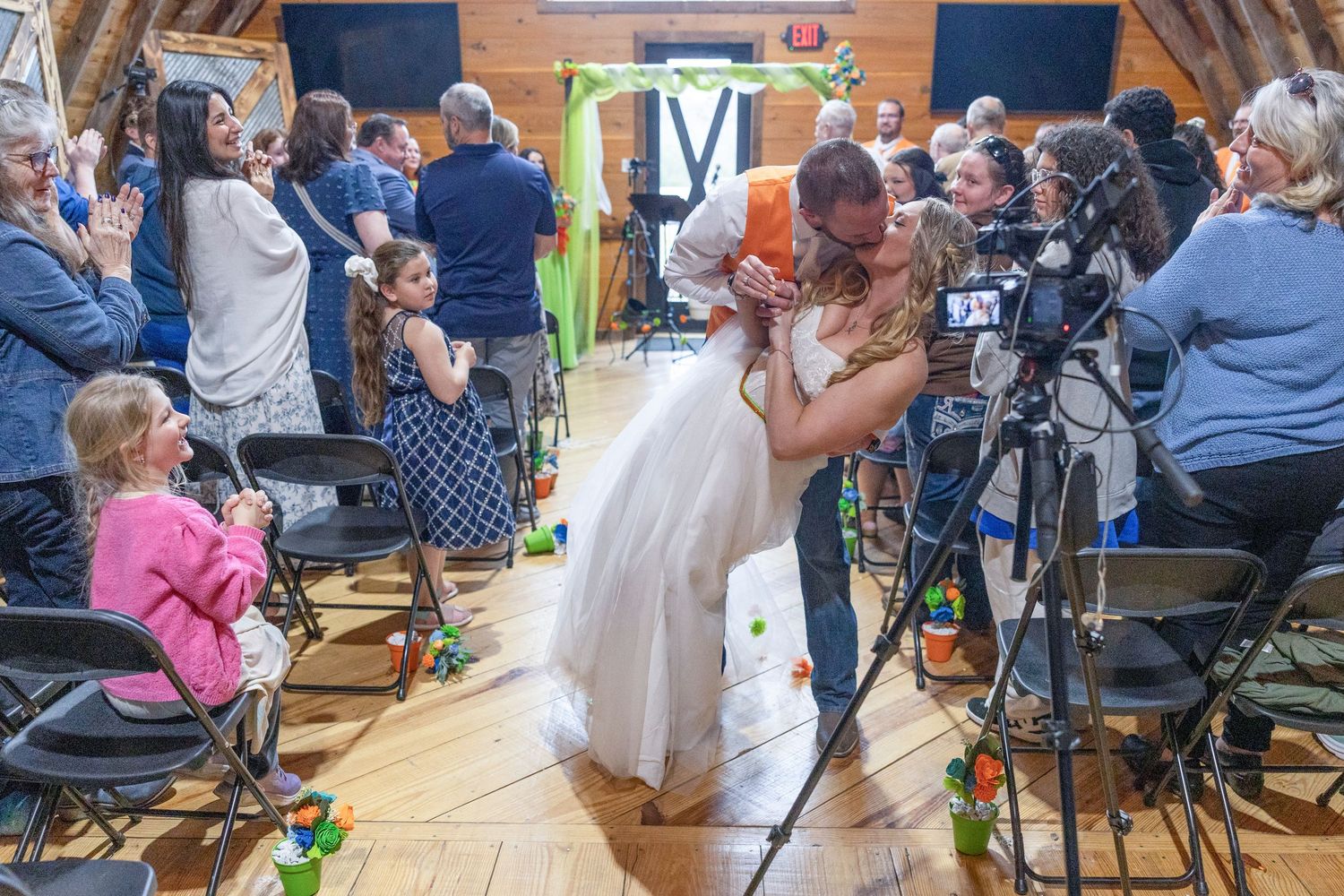 Bride and groom share first kiss at rustic barn wedding ceremony with guests applauding and videographer capturing the moment.