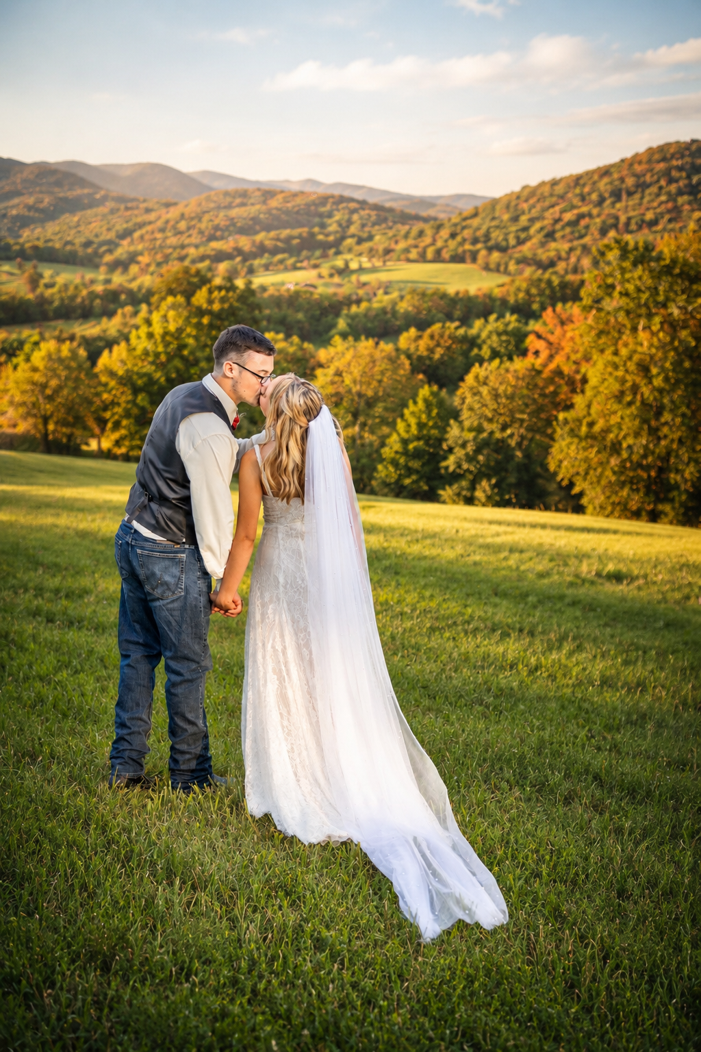 A bride and groom share a kiss in a lush green field with colorful autumn mountains at golden hour.