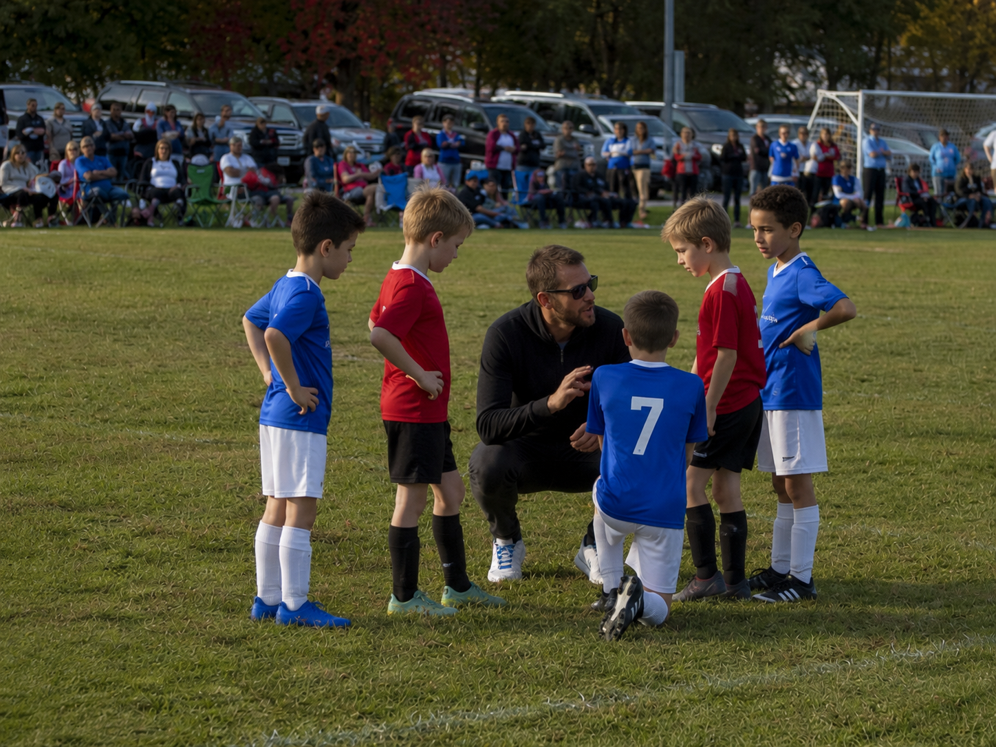 Youth soccer coach kneeling to give instructions to young players in blue and red uniforms on the field.