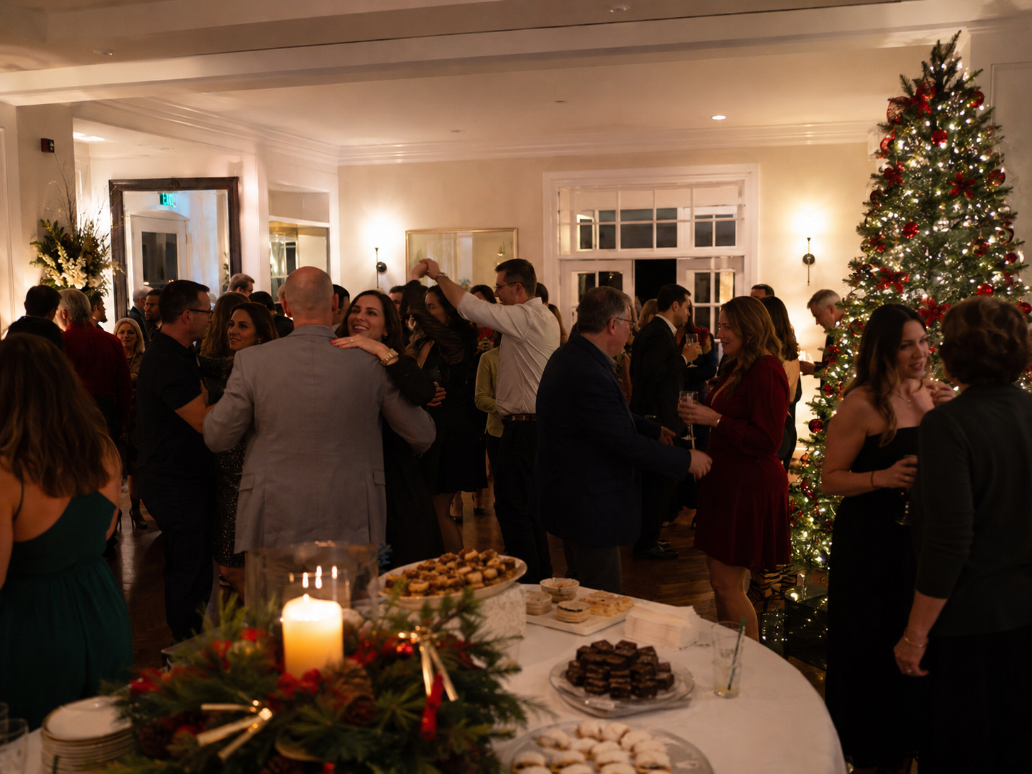 Guests dancing and socializing at a festive Christmas holiday party with decorated tree and candlelit buffet table.
