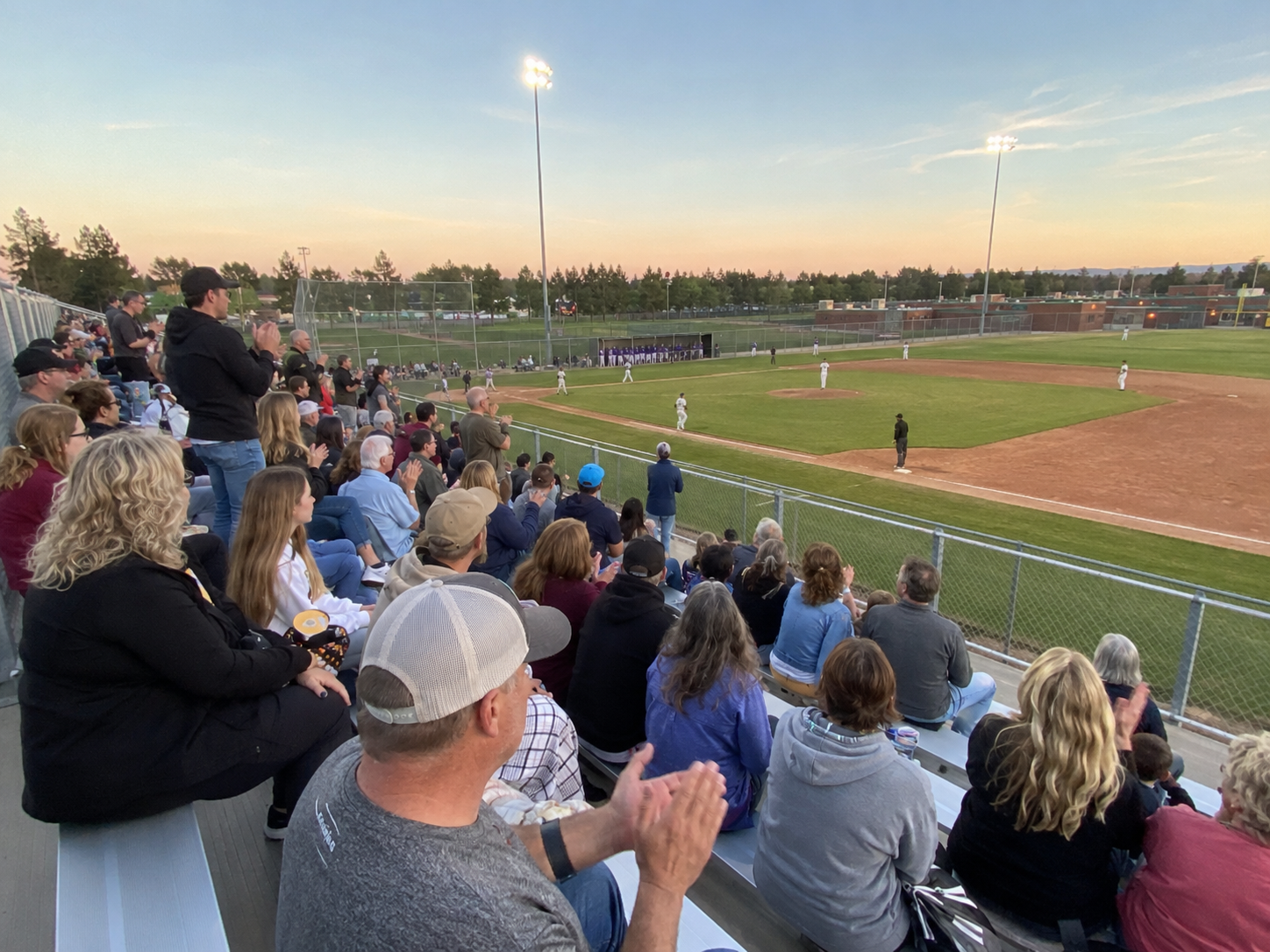 Fans stand and applaud at an outdoor baseball game during a beautiful sunset evening.