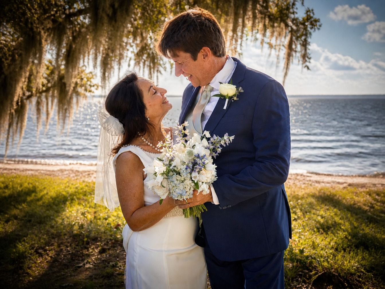 Bride and groom share a romantic moment by the waterfront, she holding a white bouquet, he in a navy suit.