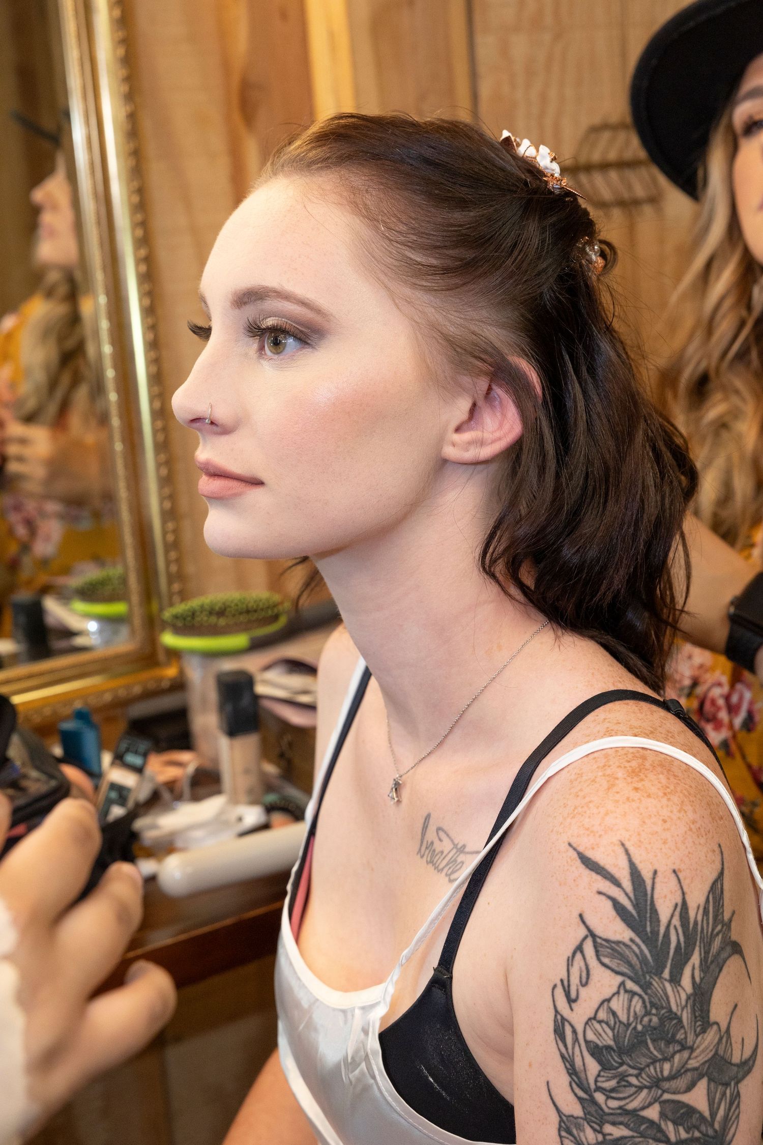 Profile view of someone getting makeup done in a dressing room, showing tattoo on shoulder.