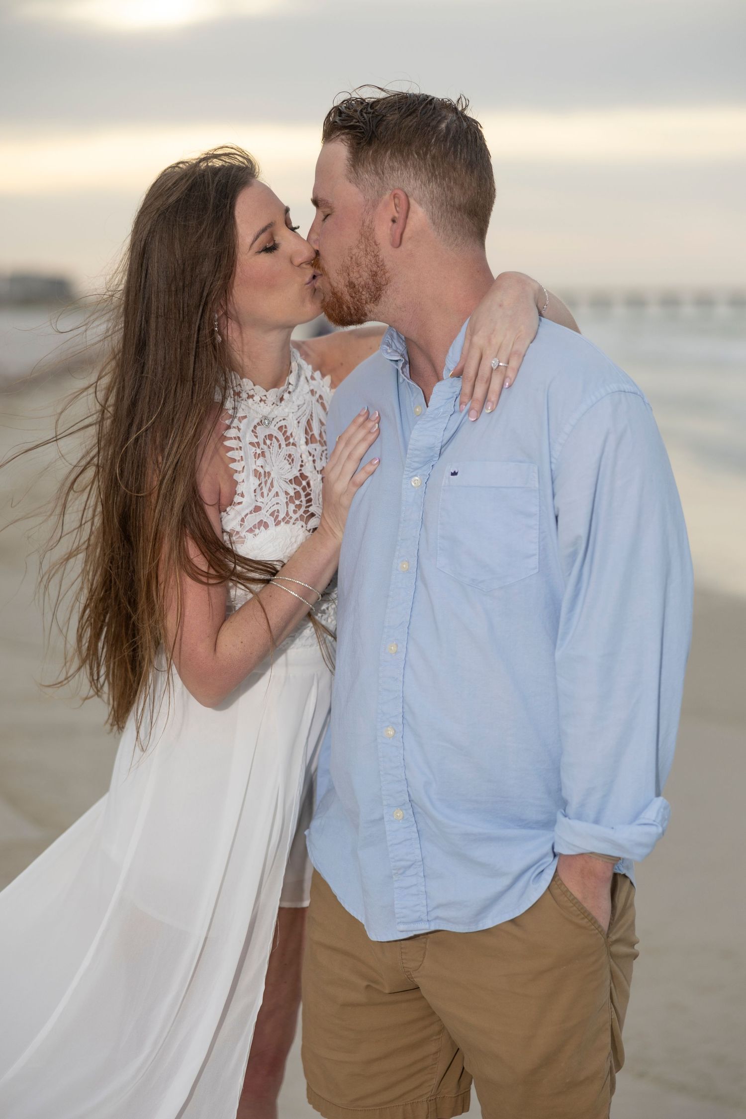 Couple sharing a romantic kiss on the beach at sunset, woman in white lace dress, man in light blue shirt.