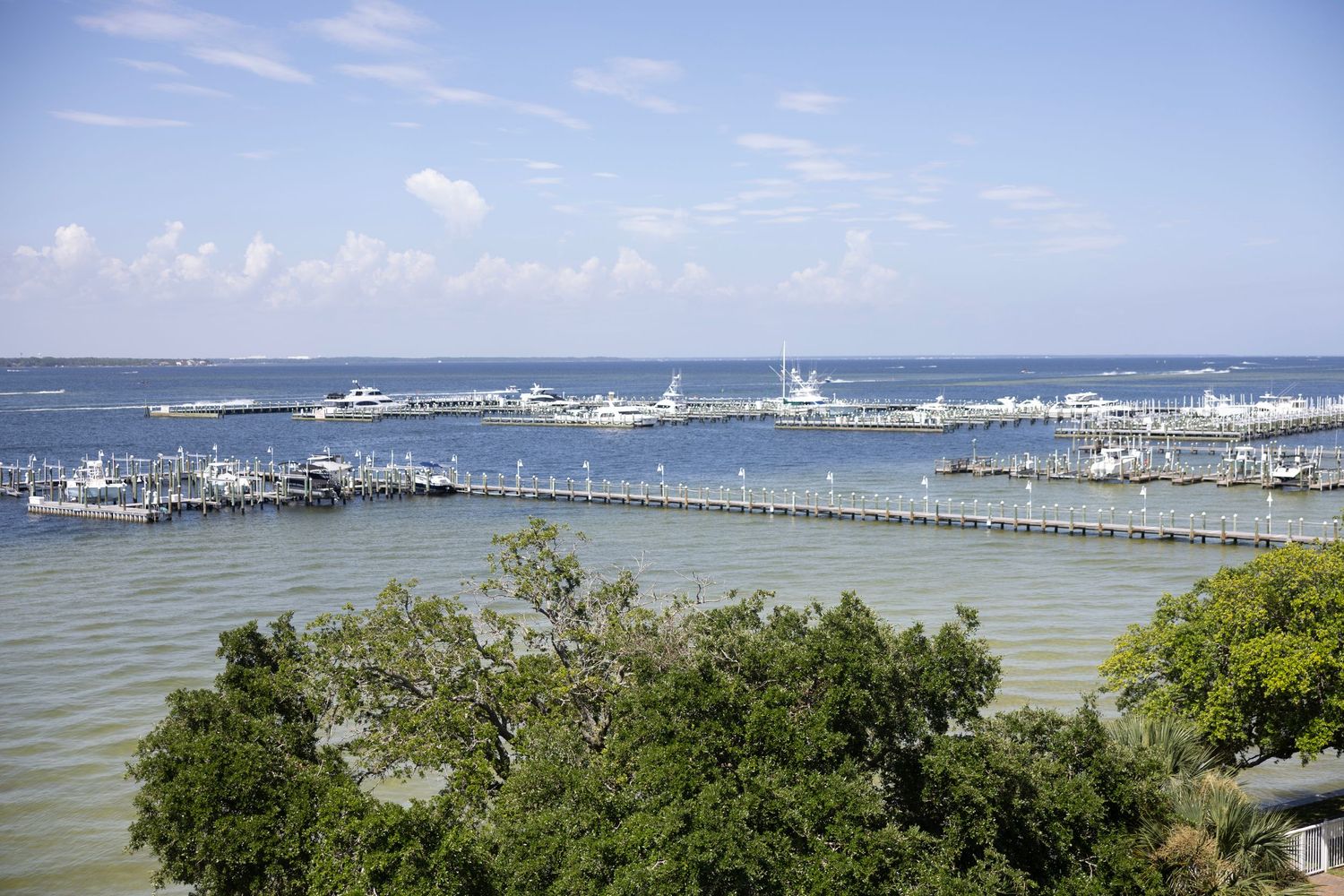 Aerial view of a marina with numerous boats docked along piers, surrounded by calm blue waters and lush green trees.