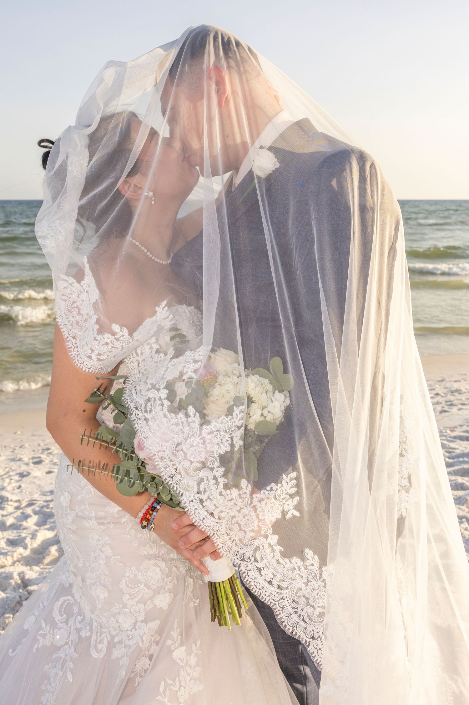 Bride and groom kissing under a flowing white veil on a beach, bride holding a white floral bouquet.