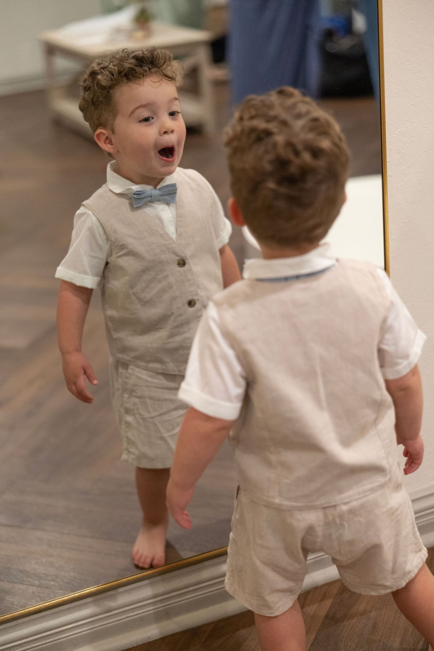 Adorable toddler boy in linen vest and bow tie gazes at his reflection in a large mirror.