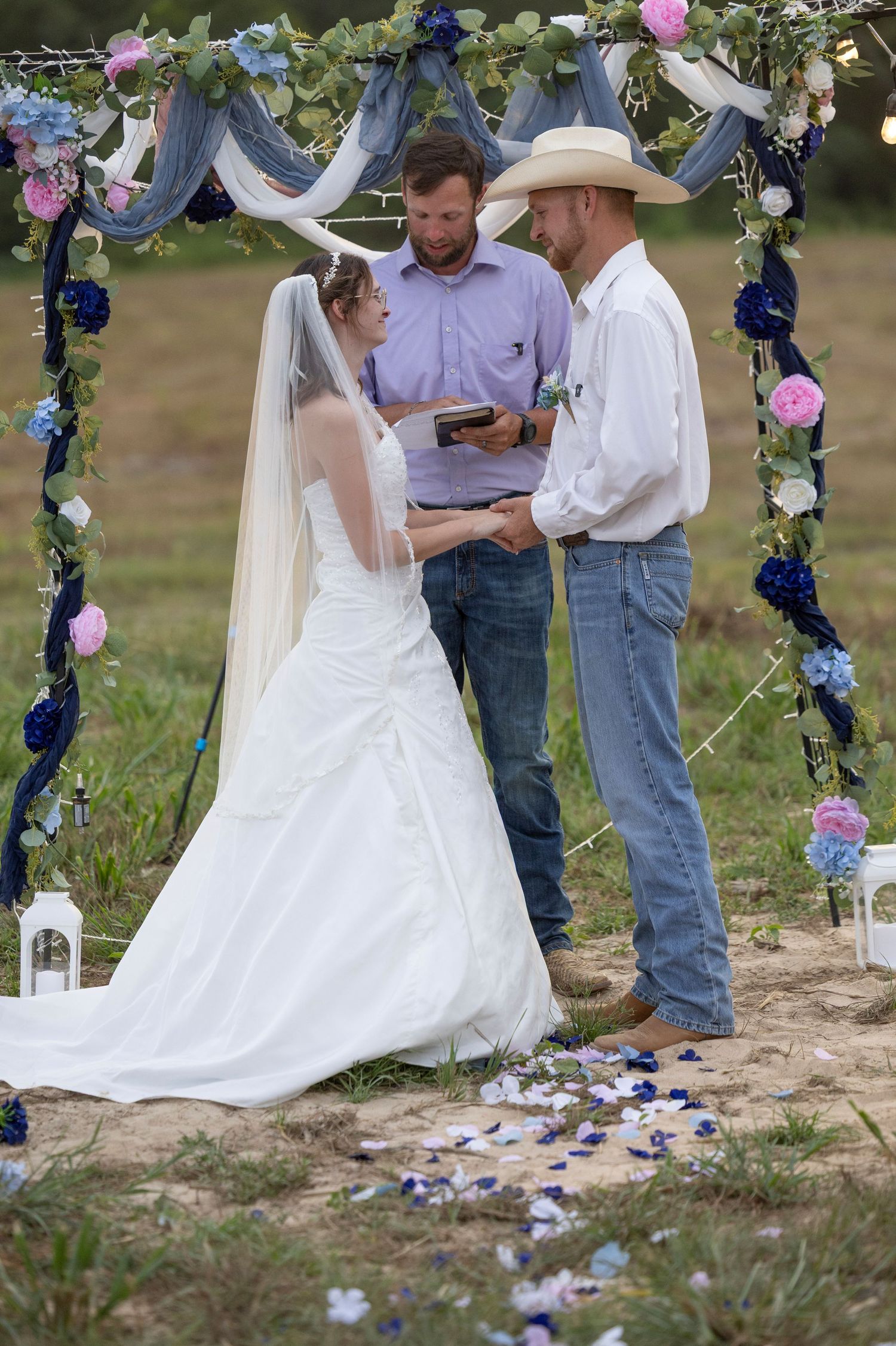 Outdoor country wedding ceremony with bride in white gown and groom in cowboy hat exchanging vows under floral arch.