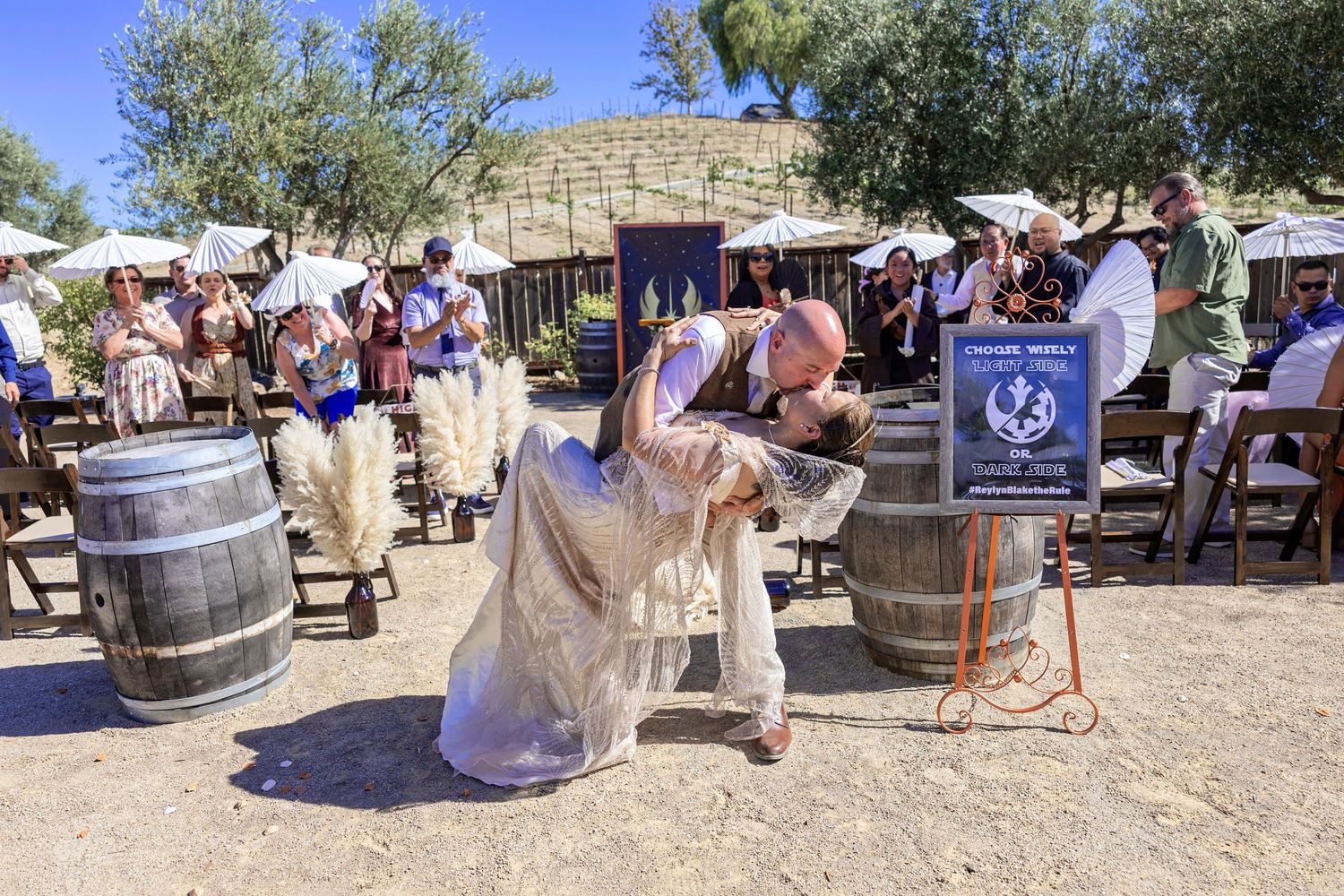 Couple shares romantic dip kiss at outdoor winery wedding ceremony with guests holding white parasols.