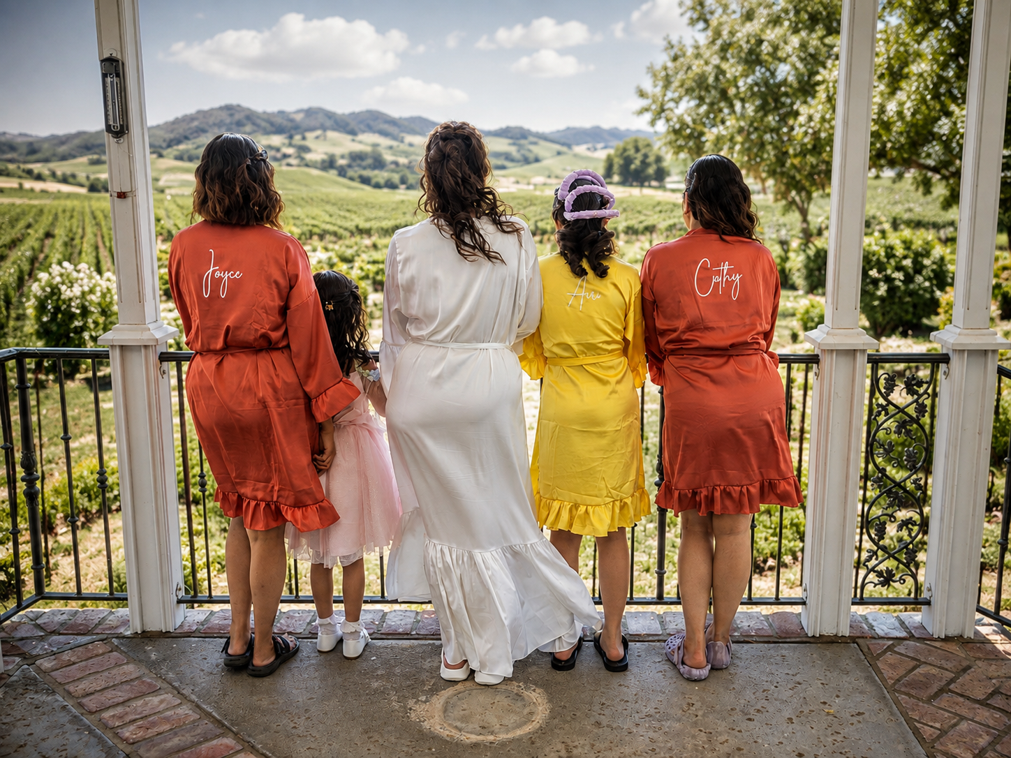 Bridesmaids in colorful robes stand on a porch overlooking lush vineyard hills with a bride in white.