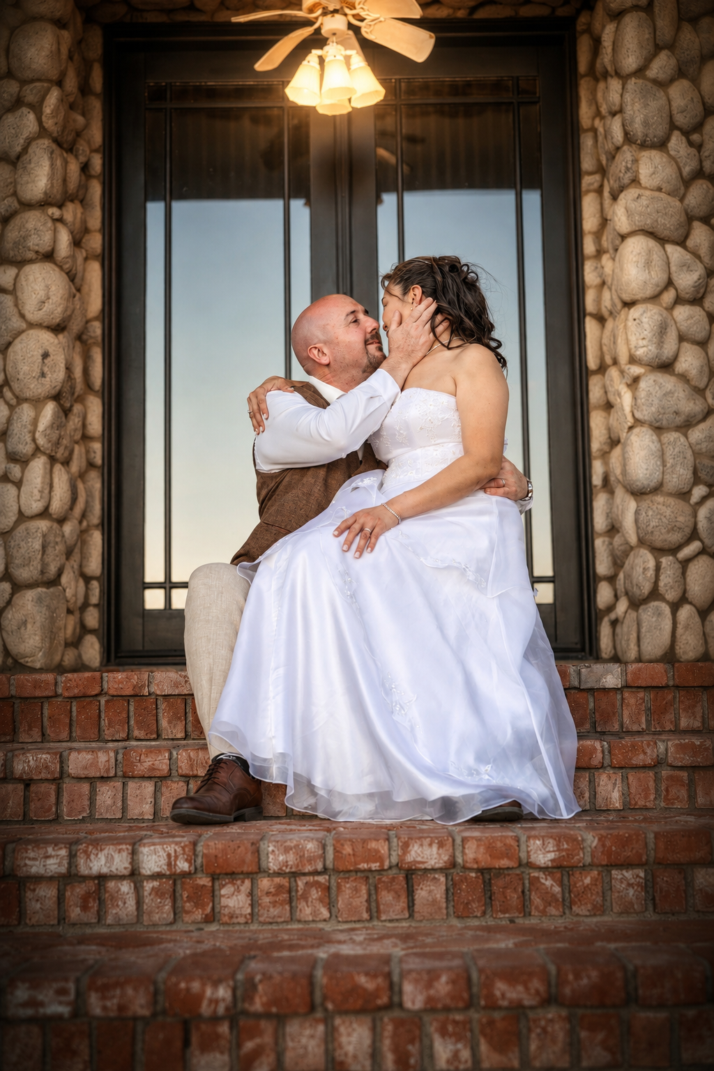 Bride and groom share a romantic kiss on brick steps outside a stone building during their wedding day.