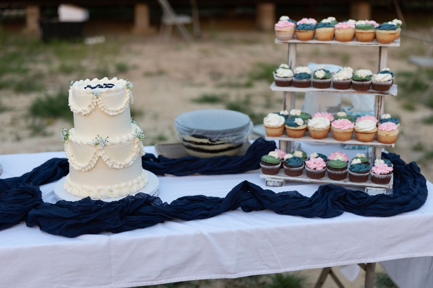 Wedding dessert table with a white tiered cake and colorful cupcakes on a stand, draped with navy blue fabric.