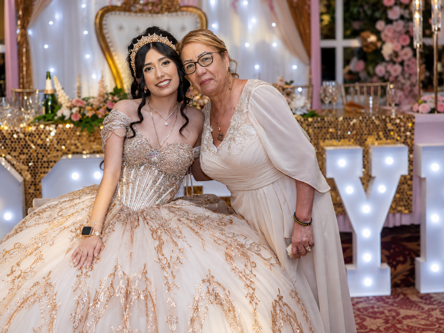 Two women pose together at a quinceañera celebration, one in an ornate gold ball gown with a tiara, surrounded by elegant decor.