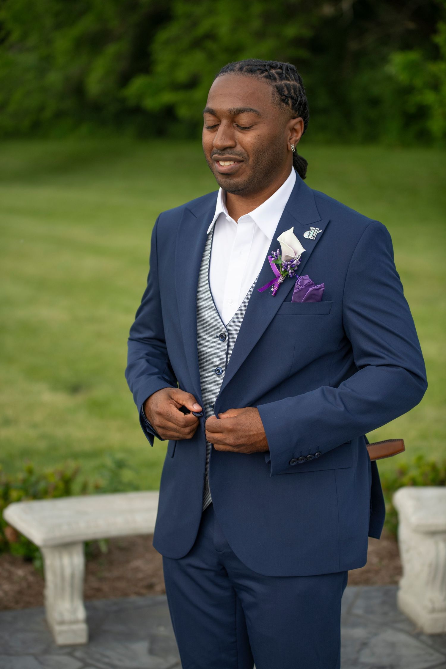 A groom in a navy blue suit and white shirt adjusts his boutonniere while standing outdoors in a garden setting.