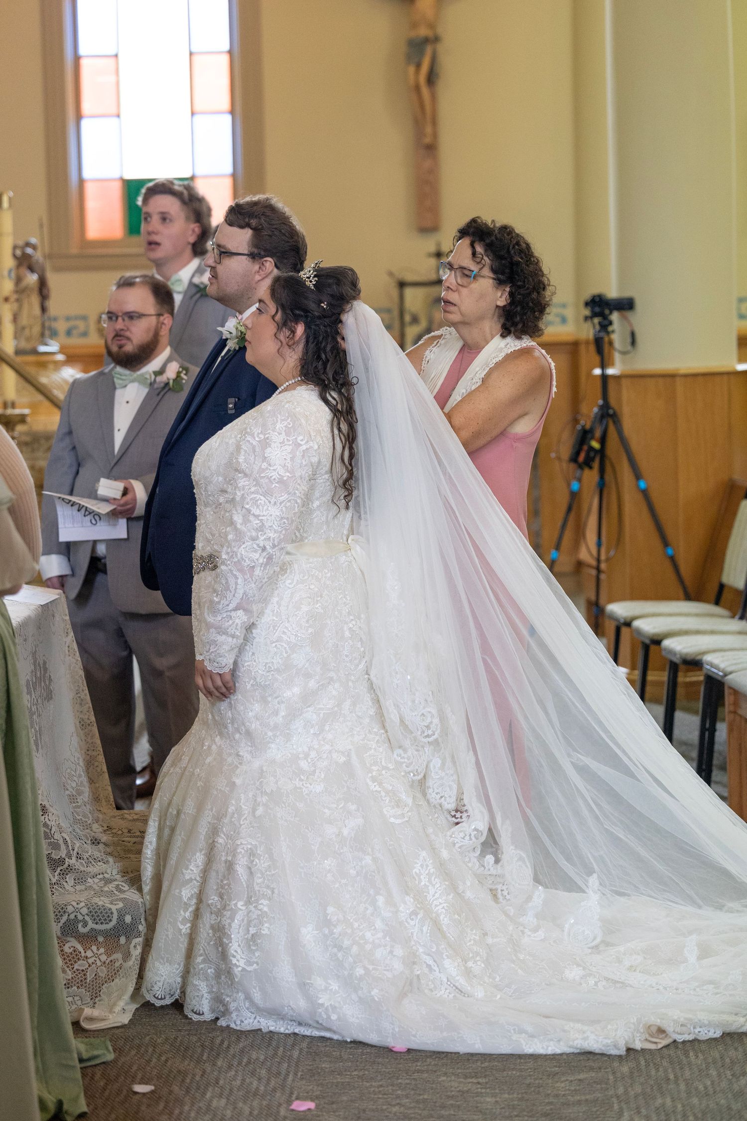 A bride in an elegant white lace wedding dress with a flowing veil stands in a church during a ceremony.