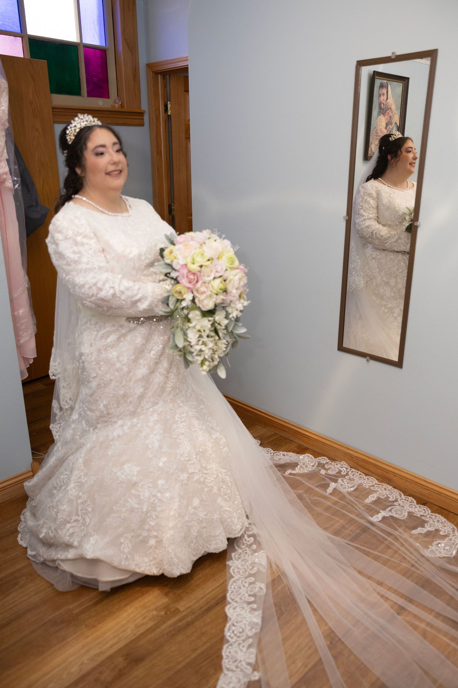 A bride in a white lace wedding dress poses with a bouquet in front of a mirror in a room with wooden floors.