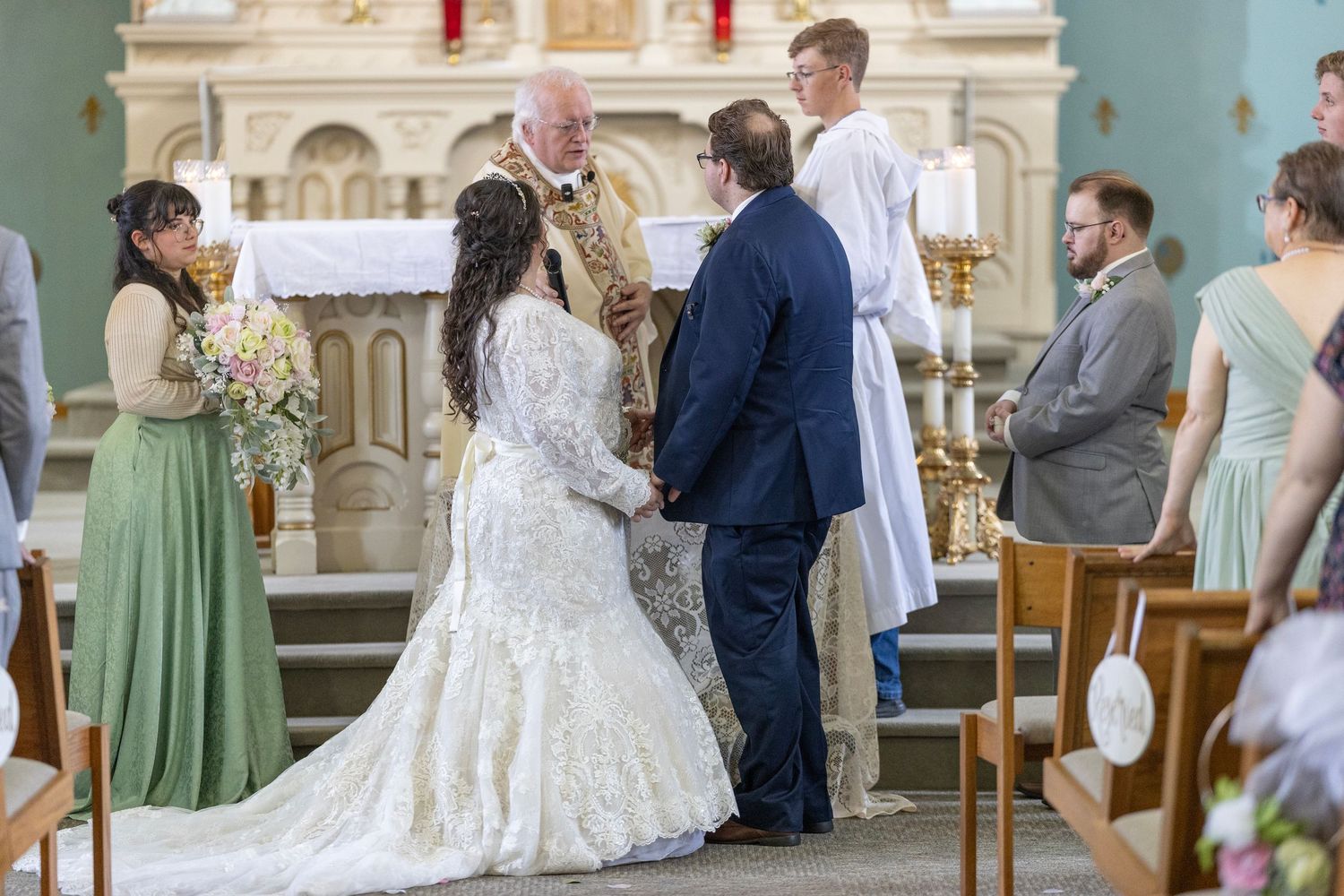 A wedding ceremony takes place in an ornate church with sage green bridesmaid dresses and white floral decorations.