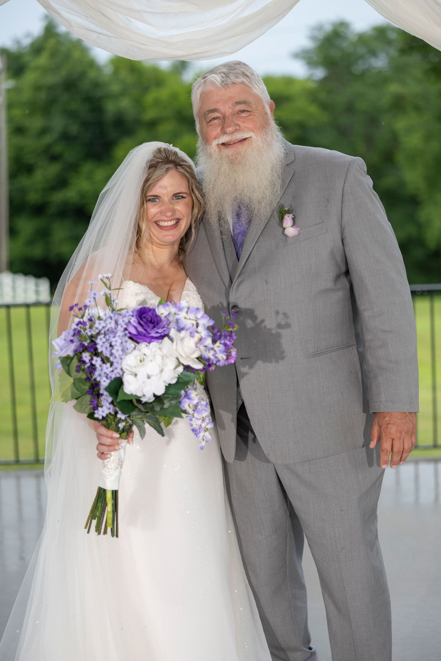 A wedding couple poses together outdoors, with the bride holding a purple and white floral bouquet and groom in gray suit.