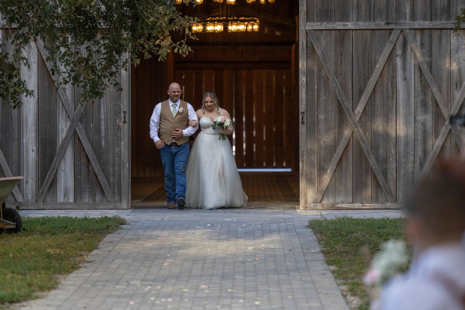 A wedding couple stands in front of a rustic barn door while their photographer captures the moment in natural light.